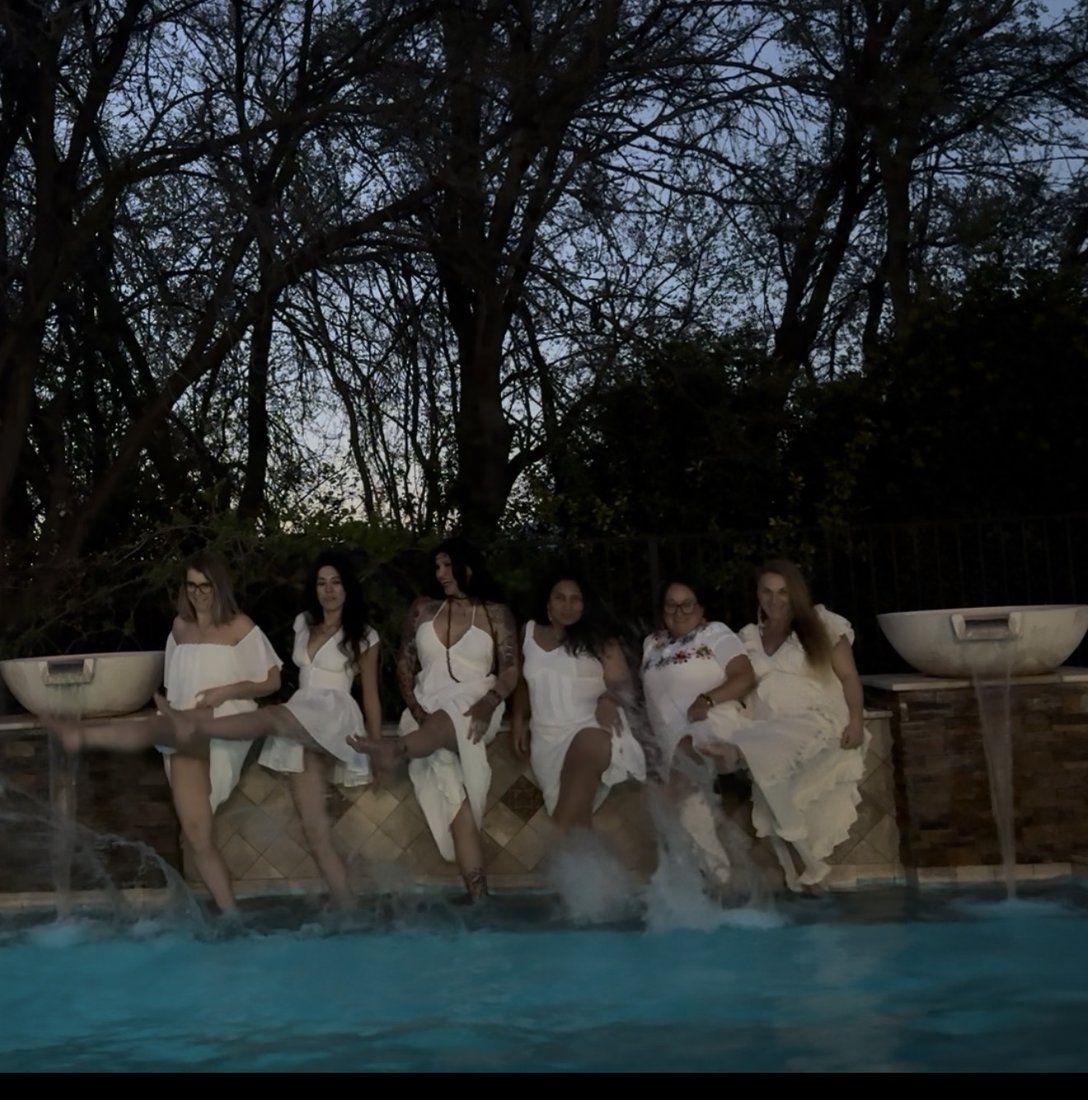 Women in white dresses splashing their feet in a glowing pool at dusk
